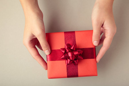 Red gift with red bow in female hands on gray background. Space for text. Top view