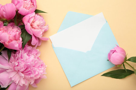 Pink peony flowers, envelope and paper card on beige background. Top view