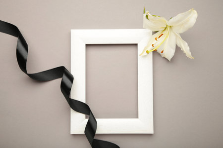 Beautiful lily flower with blank frame and black funeral ribbon on gray background.