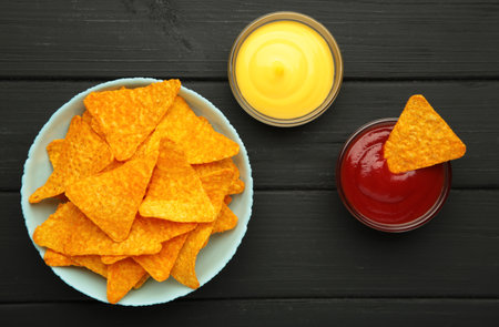 Nachos in bowl with ketchup and cheese sauce on black wooden background. Mexican dish