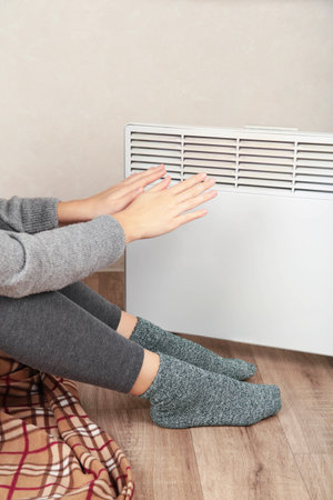 Young woman in warm socks near electric heater at home. Concept of heating season. Verticalの写真素材