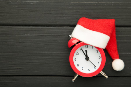 Red alarm clock with Christmas Santa hat on black wooden background. Space for text