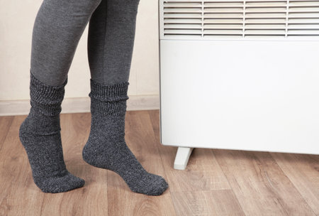 Young woman in warm socks near electric heater at home. Concept of heating seasonの写真素材