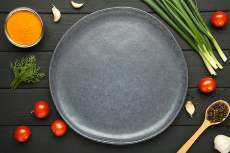 Plate with space for text and green onions, garlic, spices and tomatoes on black wooden background. Cooking concept. Top viewの写真素材