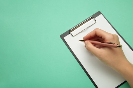 A female hand hold a pen and black clipboard with paper on mint background. Top viewの写真素材
