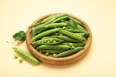 Fresh green peas in wooden bowl with pods and leaves on beige background. Top view