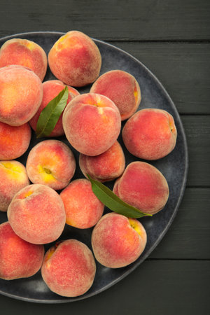Sweet peaches and leaves on dark plate on black wooden background. Top view