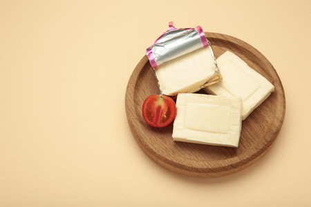 Processed cheese in foil and tomato on wooden plate on beige background. Top view
