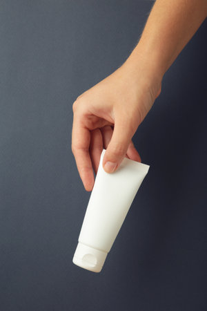 Hand holding blank white plastic tube on dark blue background. Cosmetic beauty product branding...