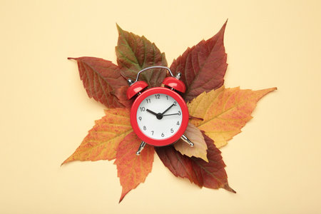 Red alarm clock, acorns and autumn leaves on beige background. Daylight saving time end. Top view