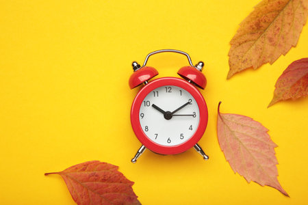 Red alarm clock, acorns and autumn leaves on yellow background. Daylight saving time end. Top view