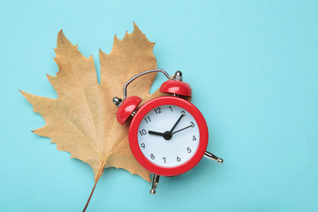 Red alarm clock and autumn leaves on blue background. Daylight saving time end. Top view