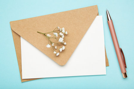 Craft envelope with paper card, pen and white flowers on pastel blue background. Top view