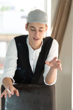 A young man in a cap and vest smokes by the windowの写真素材