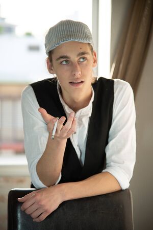 A young man in a cap and vest smokes by the windowの写真素材