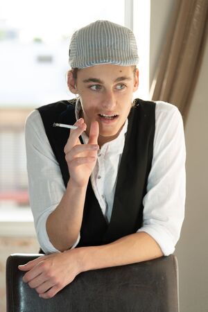 A young man in a cap and vest smokes by the windowの写真素材