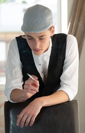 A young man in a cap and vest smokes by the windowの写真素材