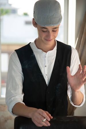 A young man in a cap and vest smokes by the windowの写真素材