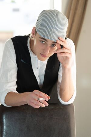 A young man in a cap and vest smokes by the windowの写真素材