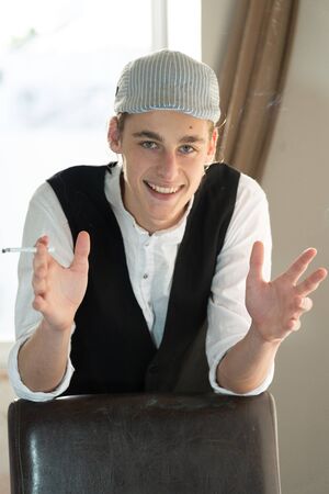 A young man in a cap and vest smokes by the windowの写真素材