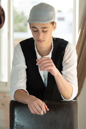 A young man in a cap and vest smokes by the windowの写真素材
