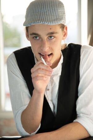 A young man in a cap and vest smokes by the windowの写真素材