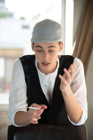 A young man in a cap and vest smokes by the windowの写真素材