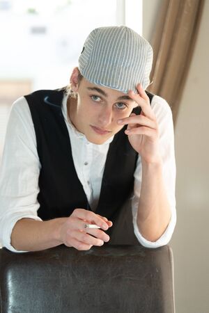 A young man in a cap and vest smokes by the windowの写真素材