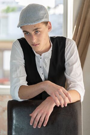 A young man in a cap and vest smokes by the windowの写真素材