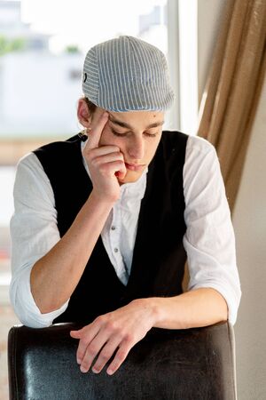 A young man in a cap and vest smokes by the windowの写真素材