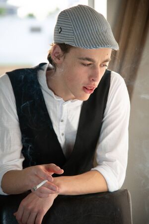 A young man in a cap and vest smokes by the windowの写真素材