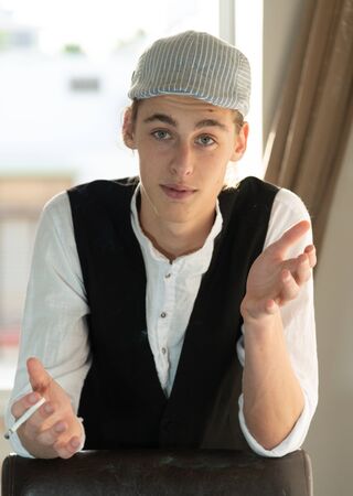 A young man in a cap and vest smokes by the windowの写真素材