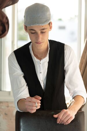A young man in a cap and vest smokes by the windowの写真素材
