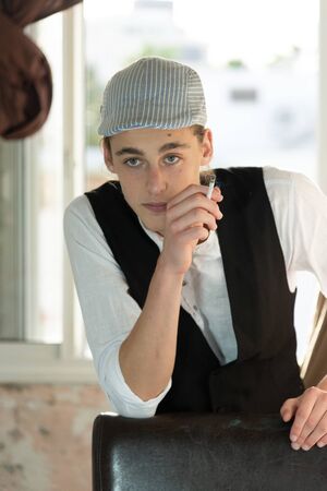 A young man in a cap and vest smokes by the windowの写真素材