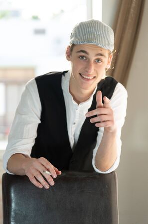 A young man in a cap and vest smokes by the windowの写真素材
