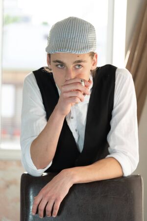 A young man in a cap and vest smokes by the windowの写真素材