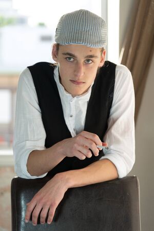 A young man in a cap and vest smokes by the windowの写真素材