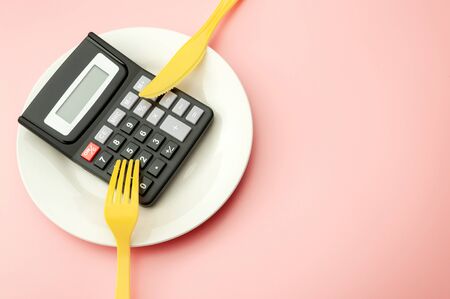 Calculate expensive food spending costs, counting calories and weight loss program concept with calculator onn empty plate, yellow fork and knife isolated on pink background with copy spaceの写真素材