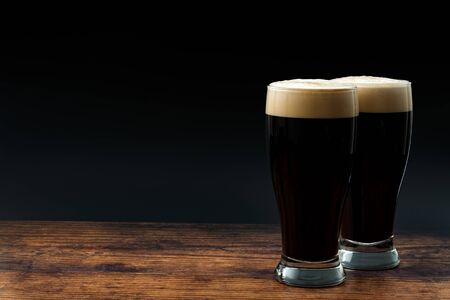 Alcohol abuse, bitter refreshing alcoholic beverage and dry stout concept theme with frothy glass pints of dark beer on wood table isolated on black background with copyspace in bar or pub settingの写真素材