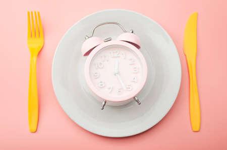 Weight loss strategy metaphor and intermittent fasting plan concept with minimal photography of clock on empty plate and vibrant yellow fork and knife isolated on vivid pink backgroundの写真素材
