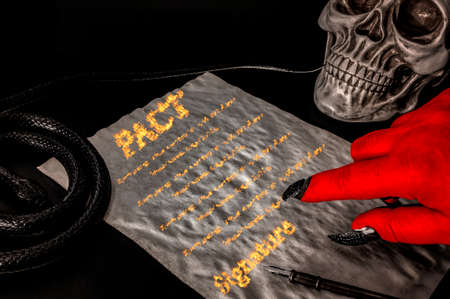 Pact or deal with the devil, faustian bargain and selling one's soul to Satan concept with red hand with claws pointing to a contract written in fire surrounded by a skull (death) and a snake (deceit)の写真素材