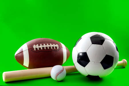 Sports equipment and leisure activity concept with a baseball bat and multiple balls used in different sports, like american football, baseball and soccer isolated on green background with copy spaceの写真素材