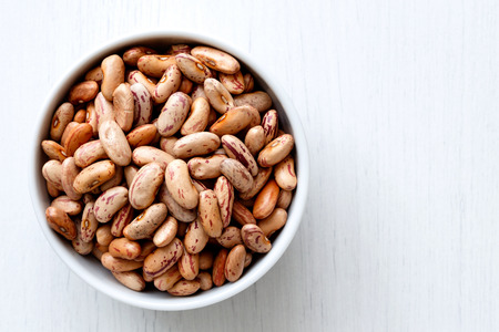Dry pinto beans in white ceramic bowl isolated on painted white wood from above.