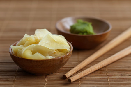 Pickled ginger slices in wooden bowl on bamboo mat next to chopsticks and wasabi paste in a bowl.の写真素材