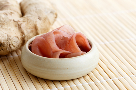 Pickled pink ginger slices in wooden bowl on bamboo mat next to ginger root.の写真素材