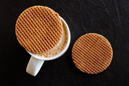 White mug with milky frothy coffee and a round waffle biscuit on top next to another waffle biscuit isolated on black leather from above.の写真素材