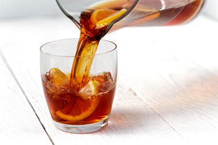 Pouring ice tea with lemons from a glass jug into a glass. White wood background.