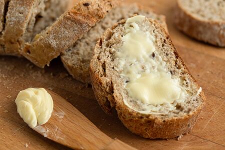Closeup of buttered slice of whole wheat rustic bread next to cut bread and wood knife. Light wood board.の写真素材