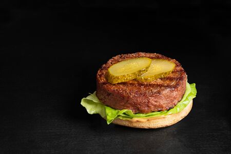 Freshly grilled plant based burger patty on bun with lettuce, slices of gherkin  and sauce isolated on black surface. Copy space.の写真素材