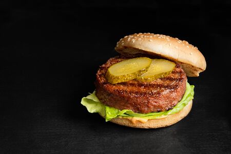 Freshly grilled plant based burger patty in bun with lettuce, slices of gherkin  and sauce isolated on black surface. Copy space.の写真素材